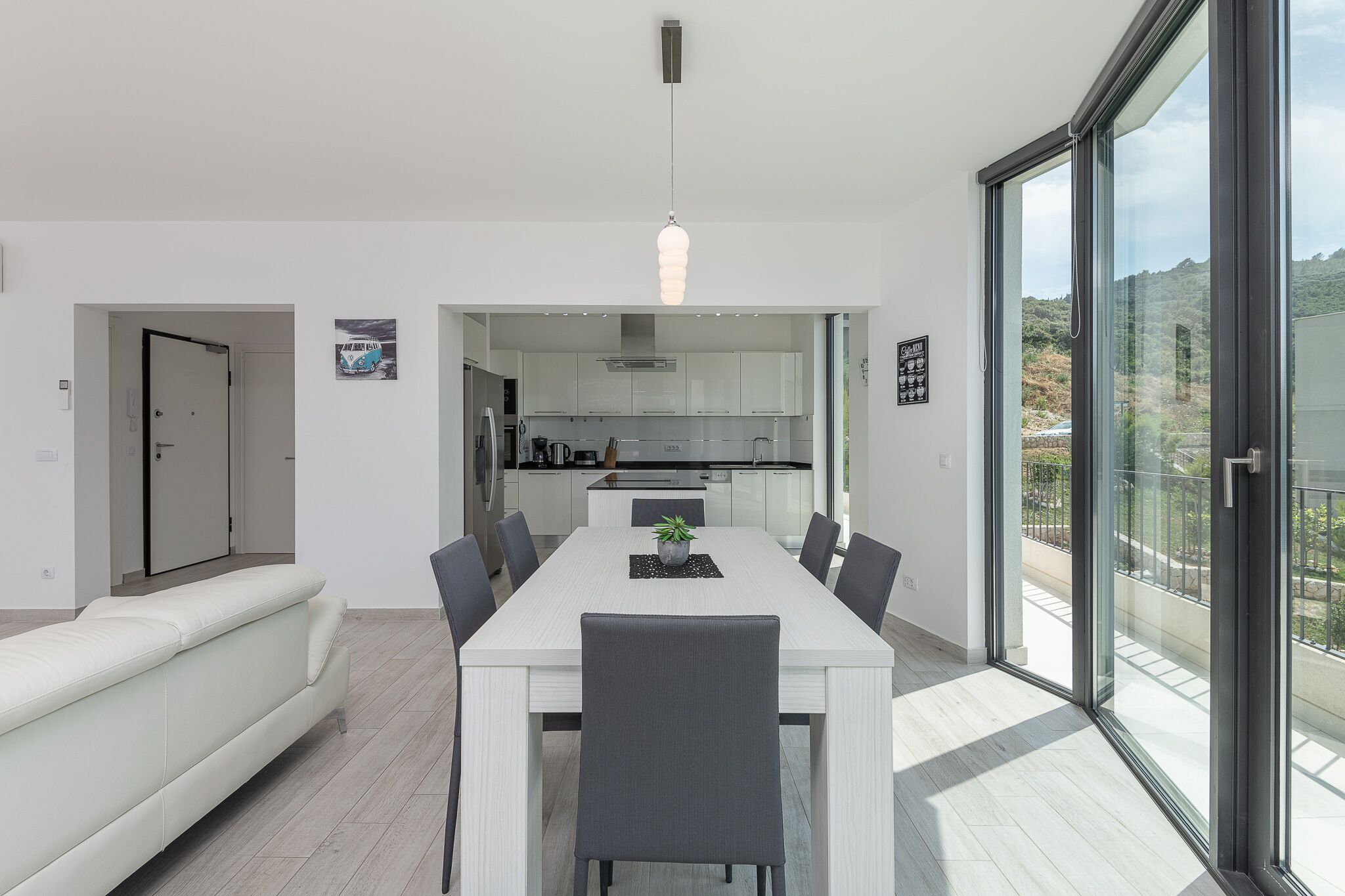 A modern dining room with a white table and chairs, a sofa, and a view of the kitchen through an open doorway.
