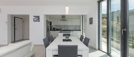 A modern dining room with a white table and chairs, a sofa, and a view of the kitchen through an open doorway.