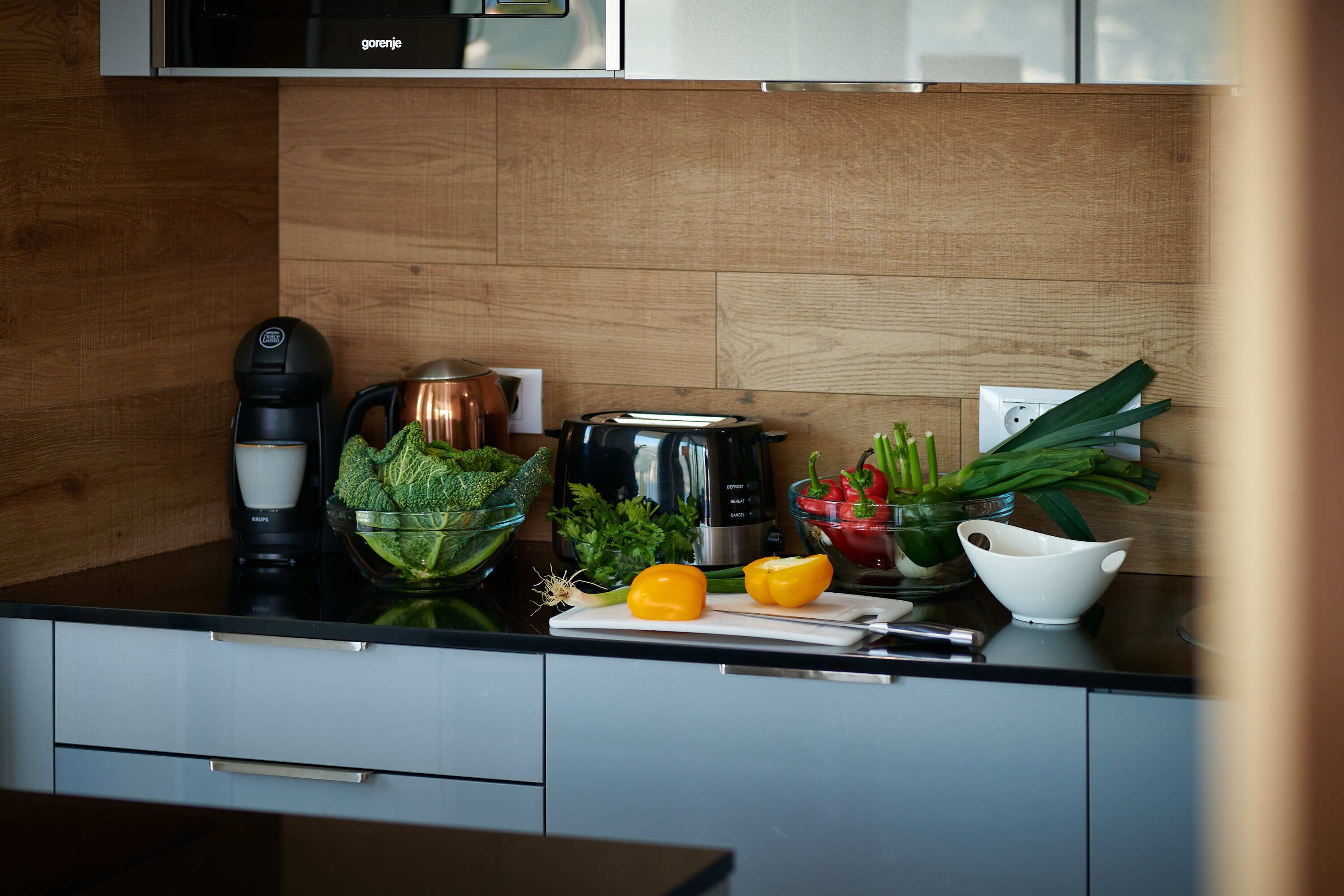 A kitchen with a coffee maker, a kettle, a toaster, and fresh vegetables on the counter.
