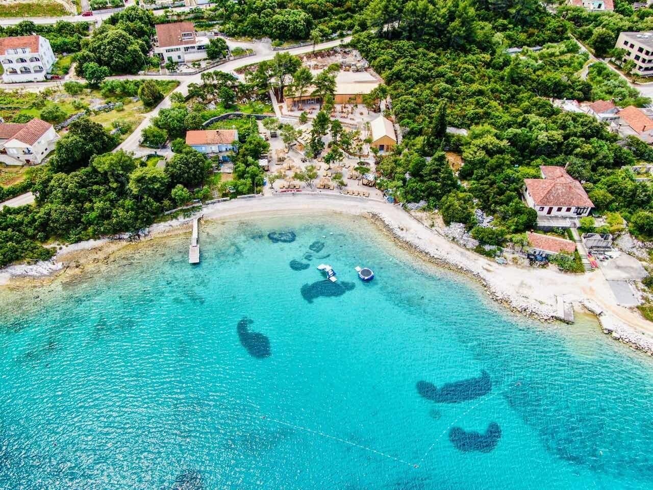 An aerial view of a coastal area with clear turquoise waters, a sandy beach, and several buildings surrounded by lush greenery.