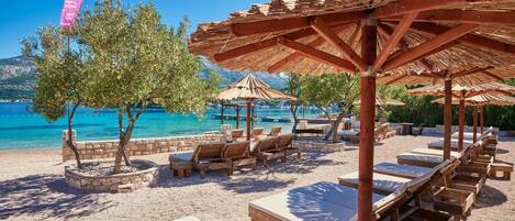 Un resort frente a la playa con sillones de madera y sombrillas de paja, una pared de piedra con árboles, y una vista despejada del mar y montañas al fondo.