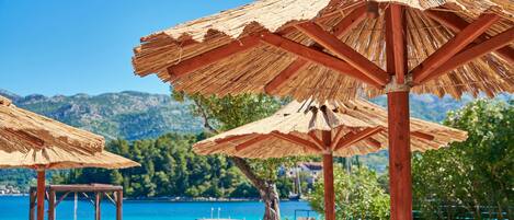 A beachside lounge with wooden chairs and thatched umbrellas, overlooking a clear blue sea and green hills.