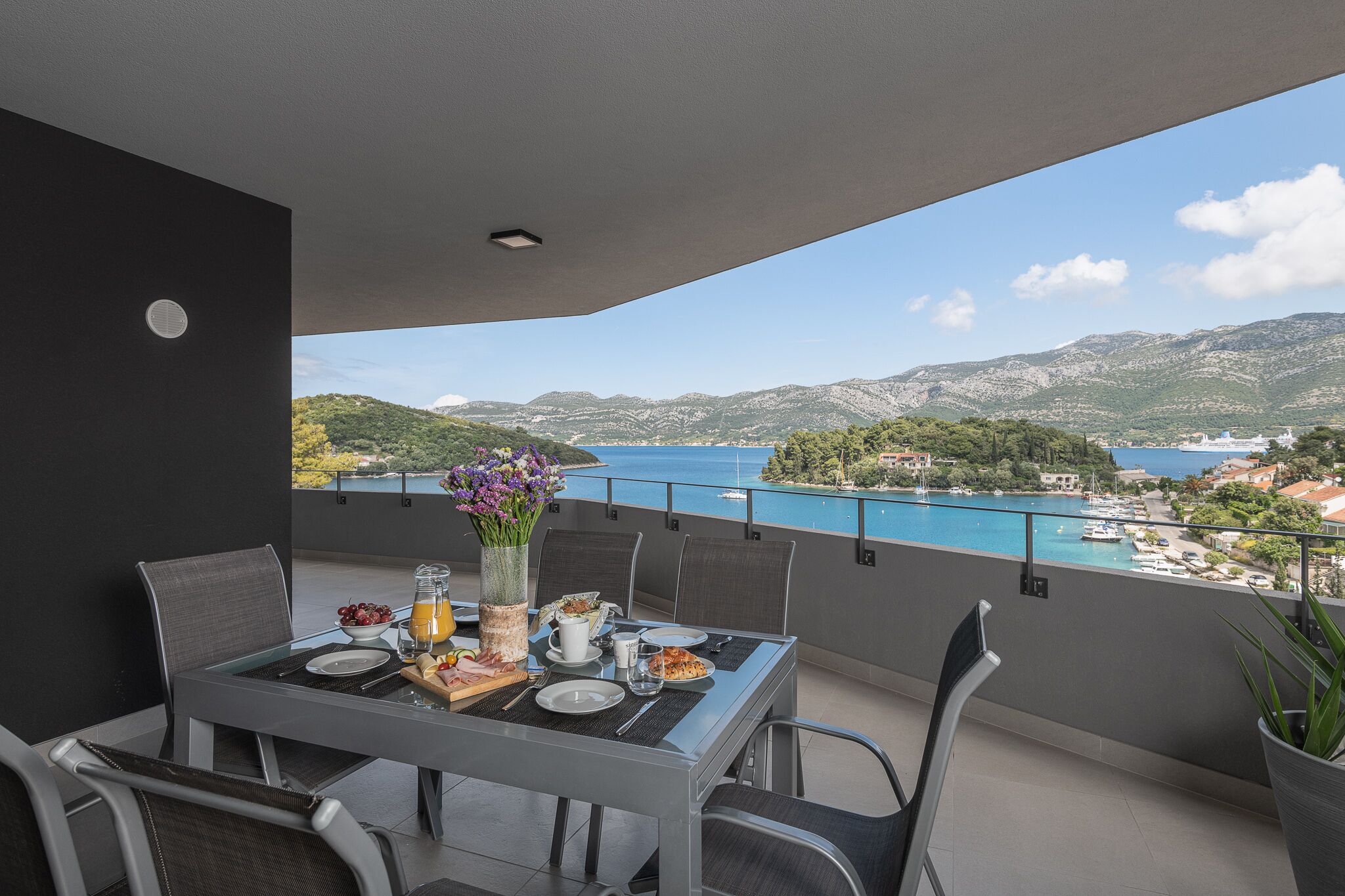 A balcony with a table set for breakfast, overlooking a scenic view of a lake and mountains.