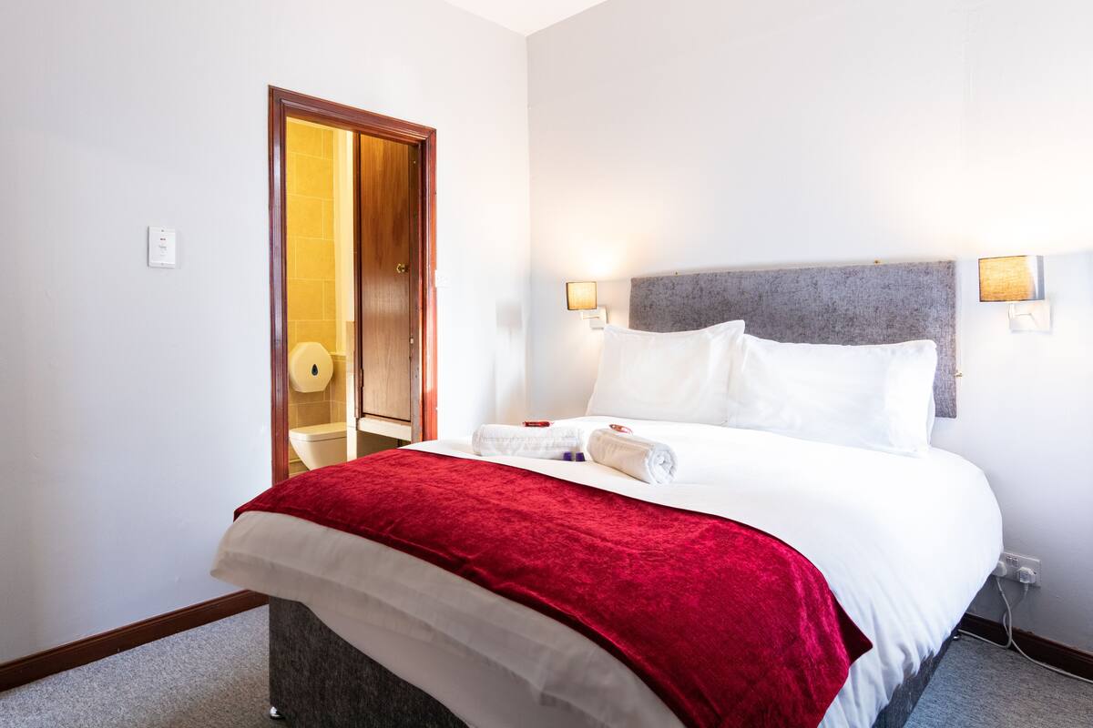 Une chambre d’hôtel avec un lit, un plaid rouge, deux lampes de chevet et une porte donnant sur une salle de bain.