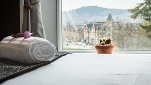 A neatly rolled white towel on a dark mat beside a window with a view of a building and mountains. - Bazpackers Hostel (Inverness)