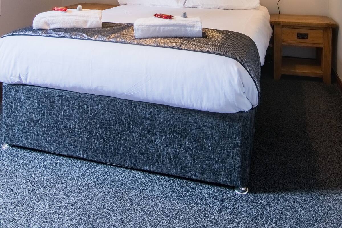 Un lit bien fait, avec du linge de lit blanc, une tête de lit sombre et une table de chevet surmontée d’une lampe.