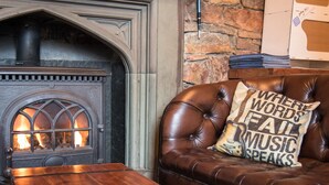 A cozy living room with a stone wall, a fireplace, a leather sofa with a cushion, and a wooden coffee table. - Bazpackers Hostel (Inverness)
