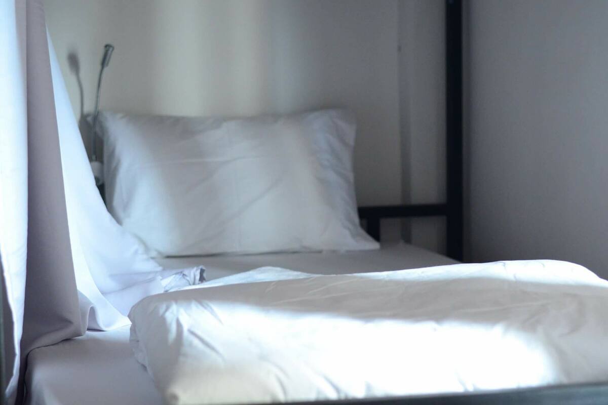 A single bed with white bedding in a room with a plain wall and a window with curtains.