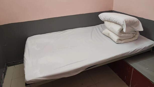A small, single-bed room with a plain white mattress and pillows, a dark headboard, and a pink wall.