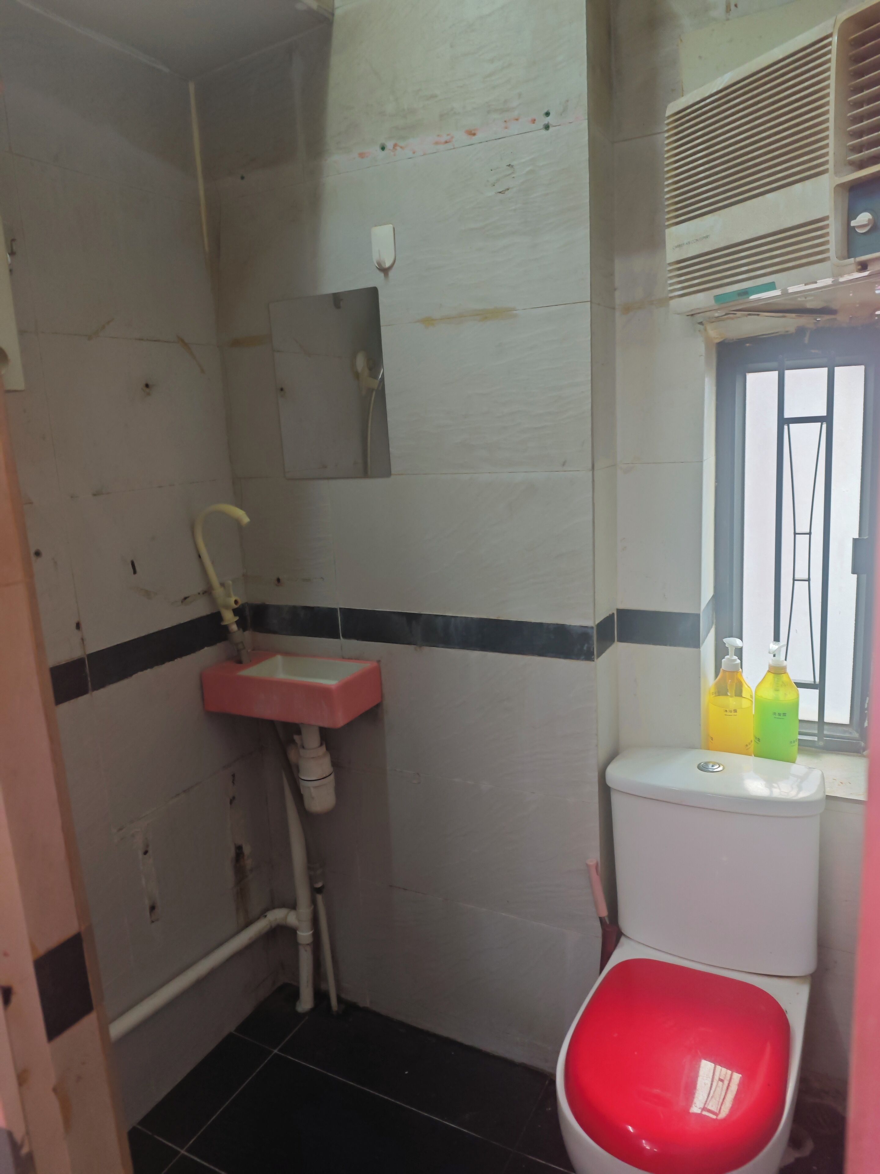A small, compact bathroom with a white toilet, a pink sink, and a window with a metal frame. There are two bottles of liquid soap on the windowsill.