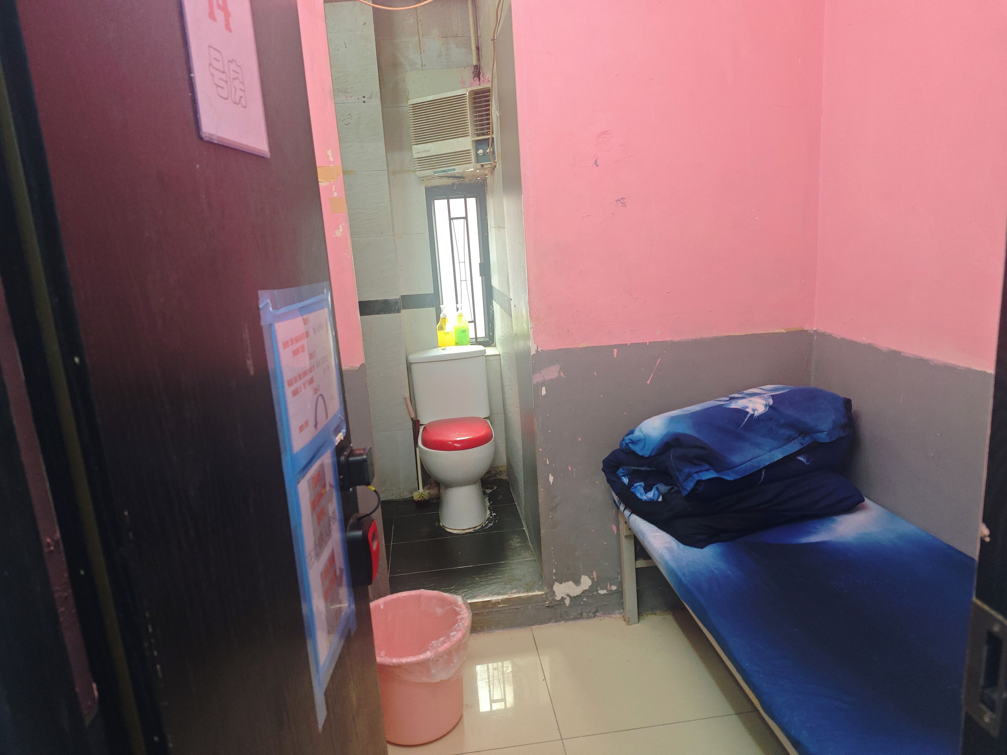 A small, narrow room with a single bed, a toilet, and a bucket.