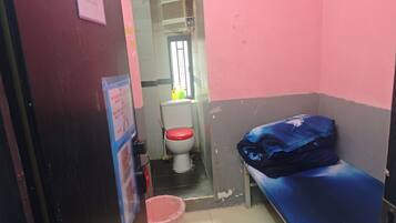 A small, narrow room with a single bed, a toilet, and a bucket.