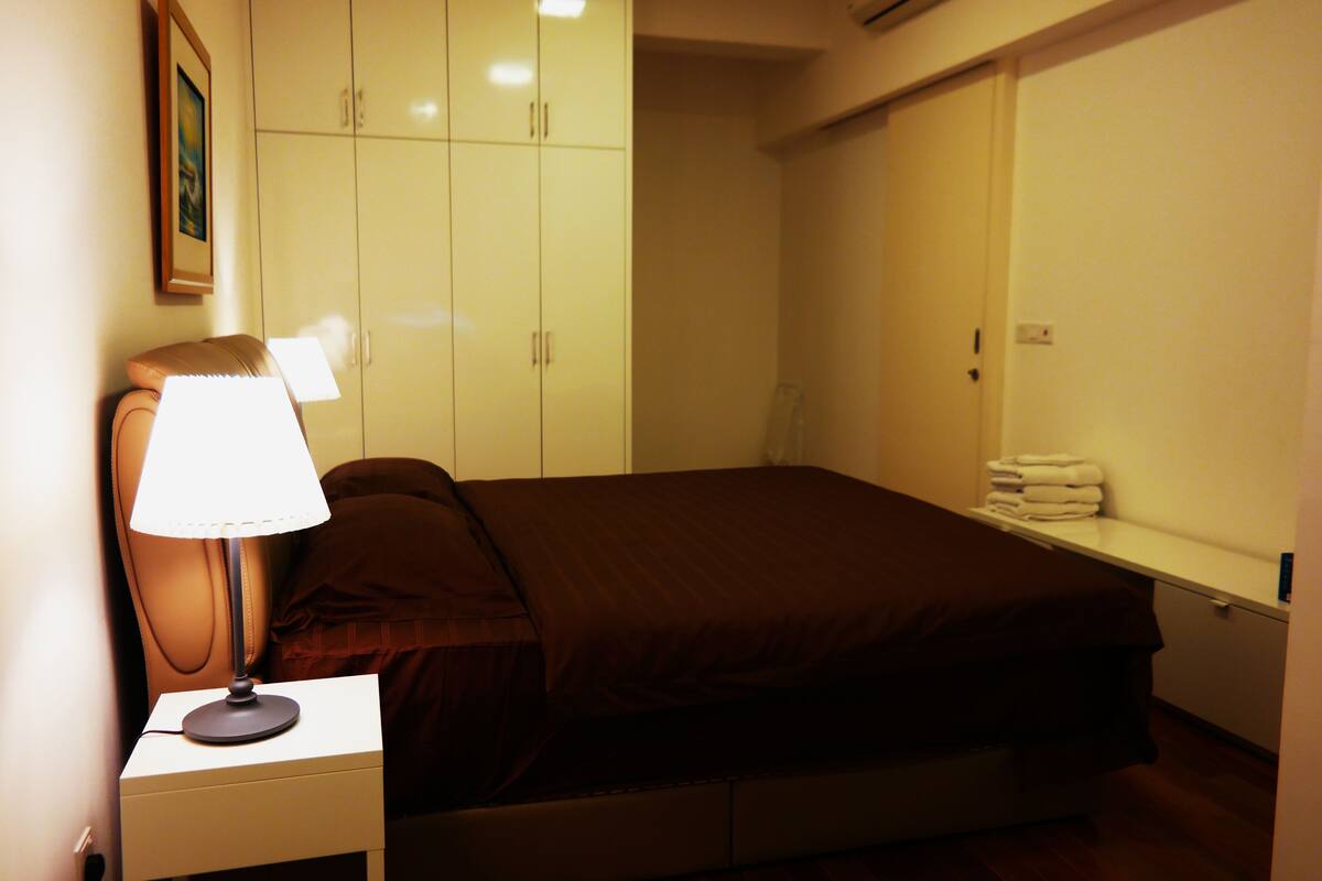 A bedroom with a bed, a nightstand with a lamp, a wardrobe, and a shelf with towels.