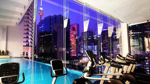 A modern hotel room with a large window overlooking a cityscape at night, featuring a swimming pool and exercise equipment.