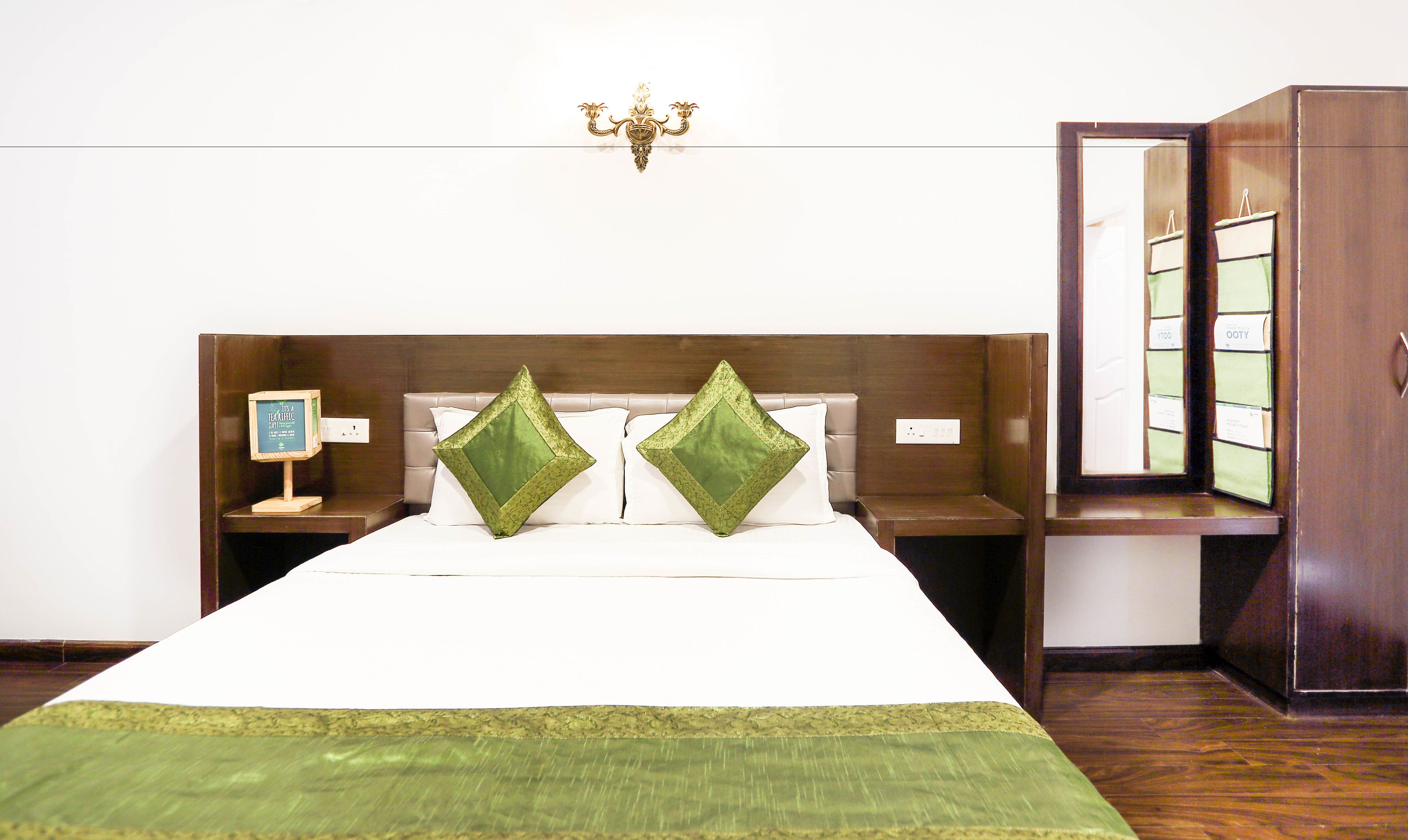 A hotel room with a large bed, two green pillows, a nightstand with a framed picture, and a mirror on the wall.