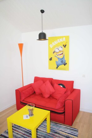 Een rode bank met meerdere kussens, een gele bijzettafel met een glas en een klein voorwerp, een zwarte hanglamp en een geel poster met een stripfiguur.
