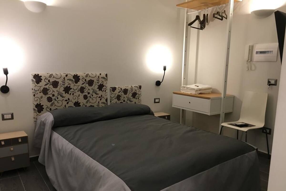 A modern bedroom with a large bed, a bedside table, a nightstand, a wall-mounted light, a mirror, and a towel rack.
