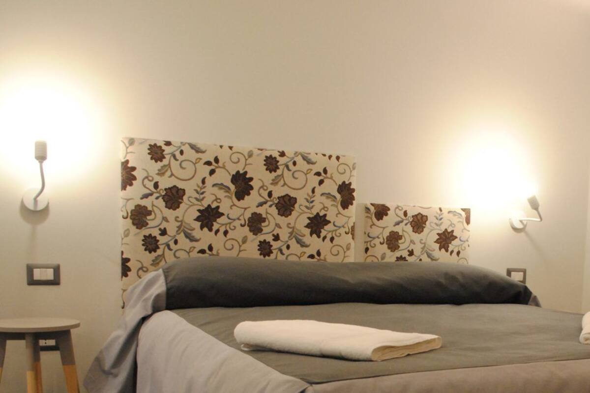 A neatly made bed with a folded towel, a wall with a floral pattern, and two wall-mounted lights.