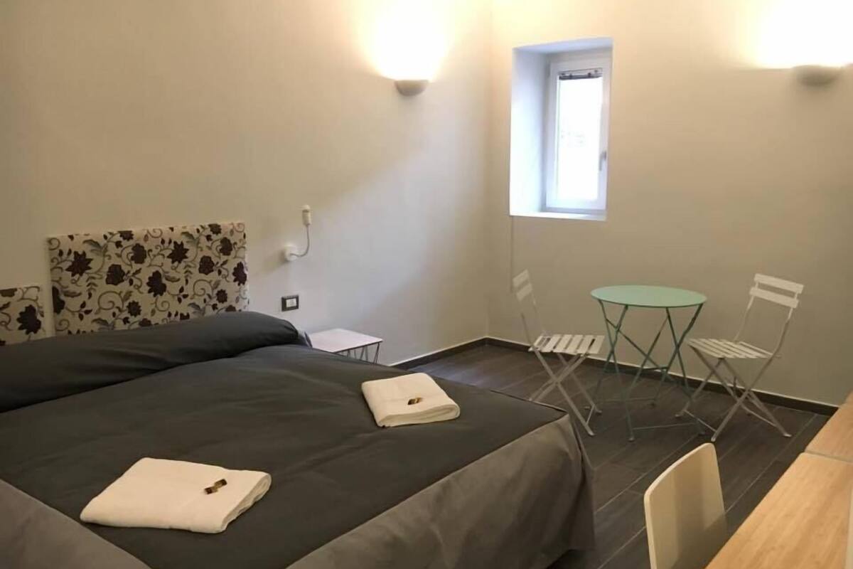 A small, well-lit room with a bed, a small table, two chairs, and a window.