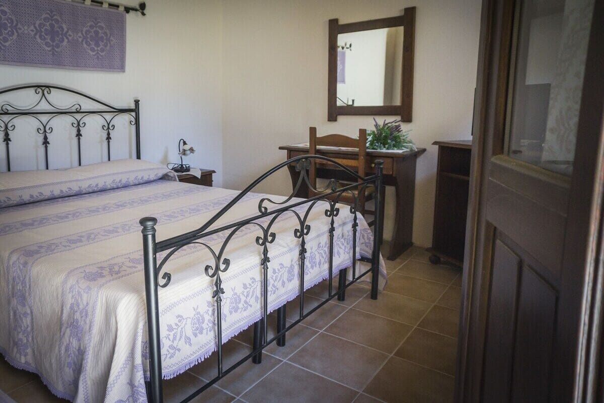A bedroom with a bed, a nightstand, a desk with a chair, a mirror, and a window.