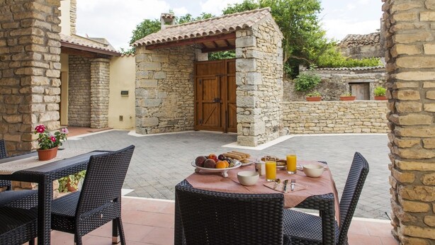 An outdoor patio with stone walls, a wooden door, wicker furniture, and a table set with fruits, cookies, and orange juice. - Albergo Diffuso Il Mandorlo (Baressa)