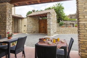 An outdoor patio with stone walls, a wooden door, wicker furniture, and a table set with fruits, cookies, and orange juice. - Albergo Diffuso Il Mandorlo (Baressa)