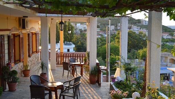 A covered outdoor seating area with stone flooring, potted plants, and a view of a hillside town. - Sofia (Apokoronas)