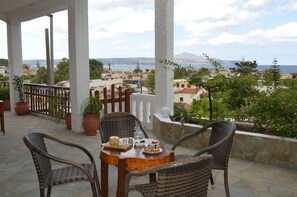 A covered outdoor seating area with wicker chairs and a table, overlooking a coastal town and a mountain. - Sofia (Apokoronas)