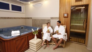 A spa room with a hot tub, two people in robes, a massage table, and a wooden wall. - Hotel Lafonte (Karlovy Vary)