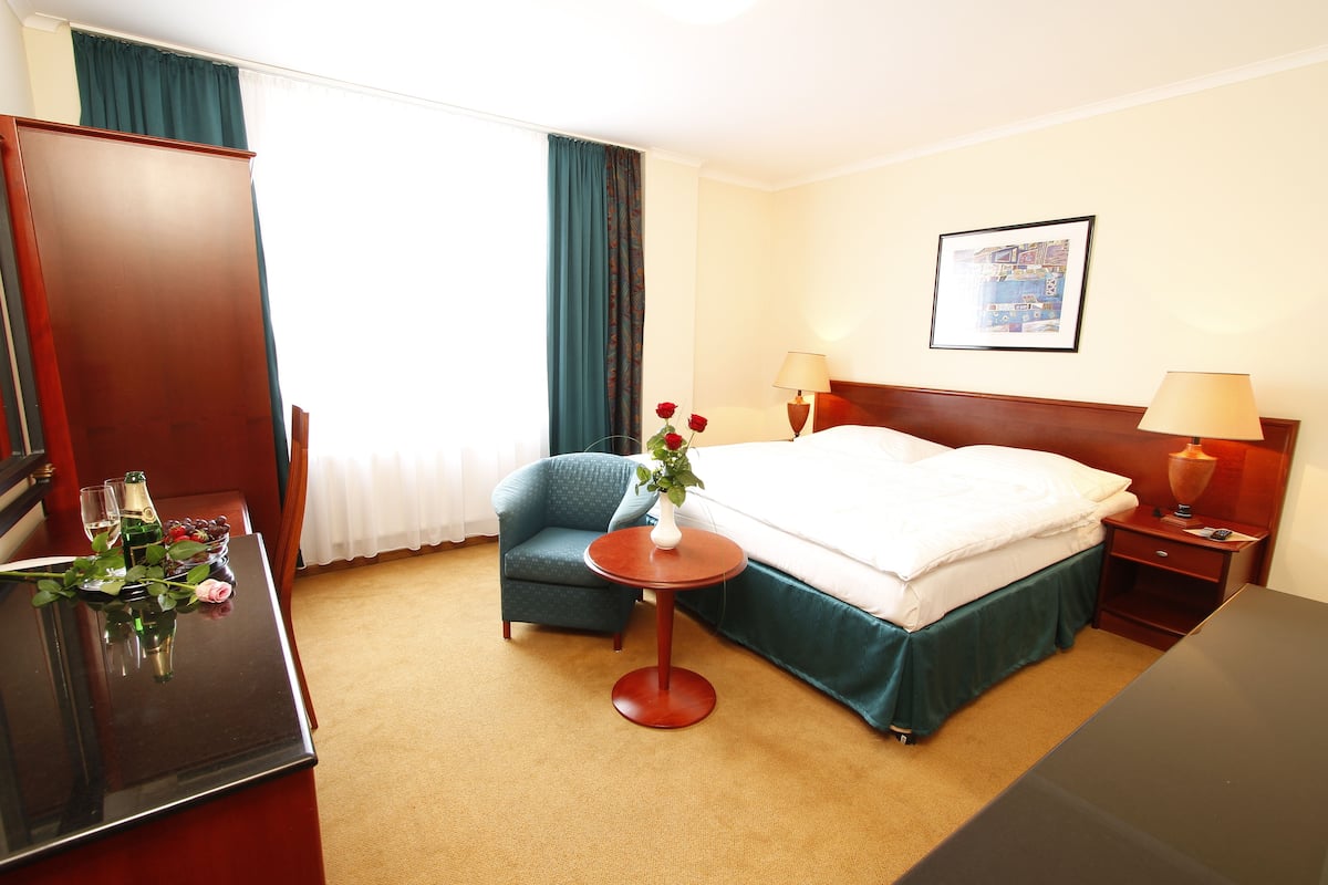 Une chambre d’hôtel avec un lit, un bureau agrémenté d’un vase de fleurs, une chaise, une table de chevet avec une lampe et un tableau encadré au mur.