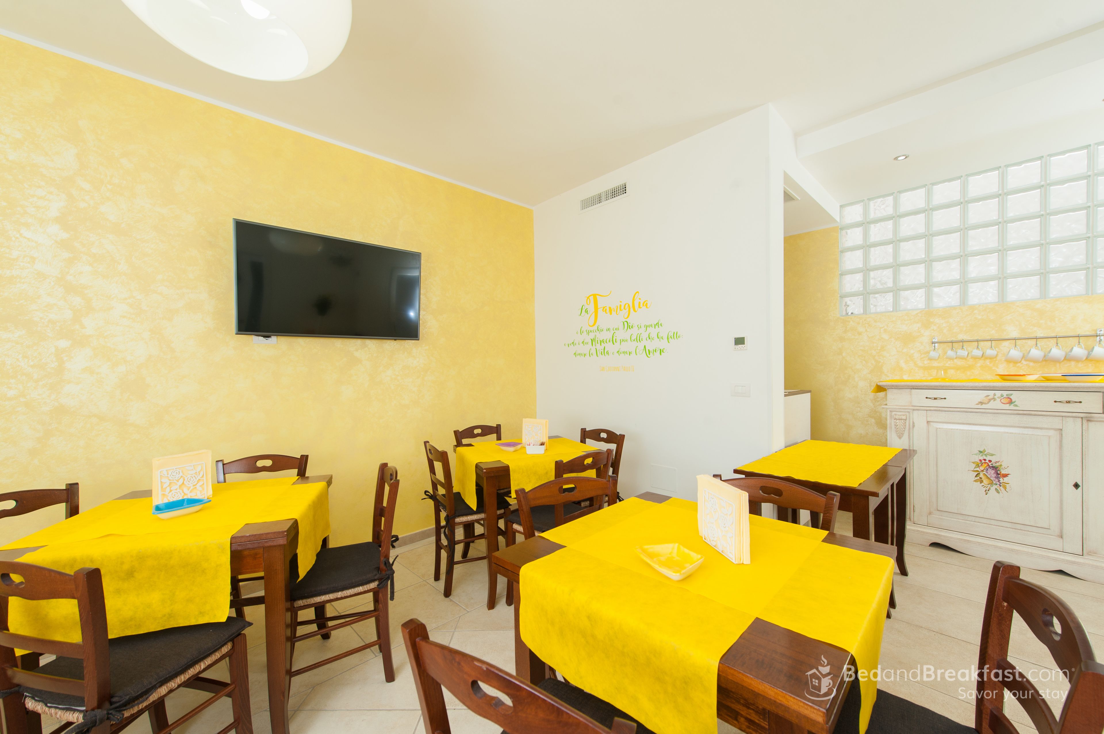 A dining area with yellow tables and chairs, a flat-screen TV mounted on the wall, and a decorative wall with text.