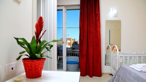 Een hotelkamer met een balkon, een rood gordijn met patroon, een wit bed en een rieten stoel.