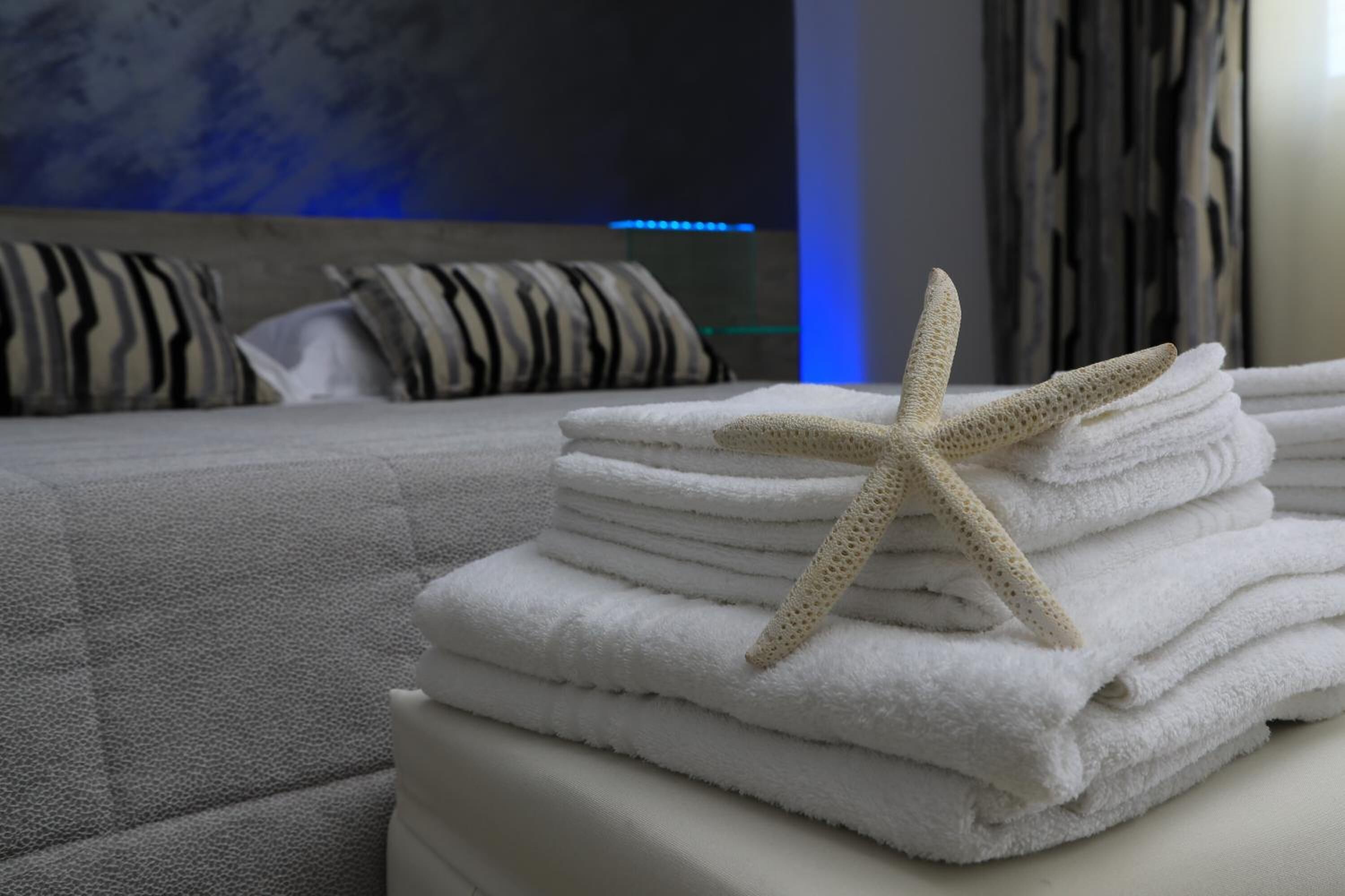 Une pile de serviettes blanches, soigneusement rangées, avec une étoile de mer posée sur le dessus, est placée sur une surface blanche dans une pièce où se trouvent un canapé gris et des coussins à rayures en arrière-plan.