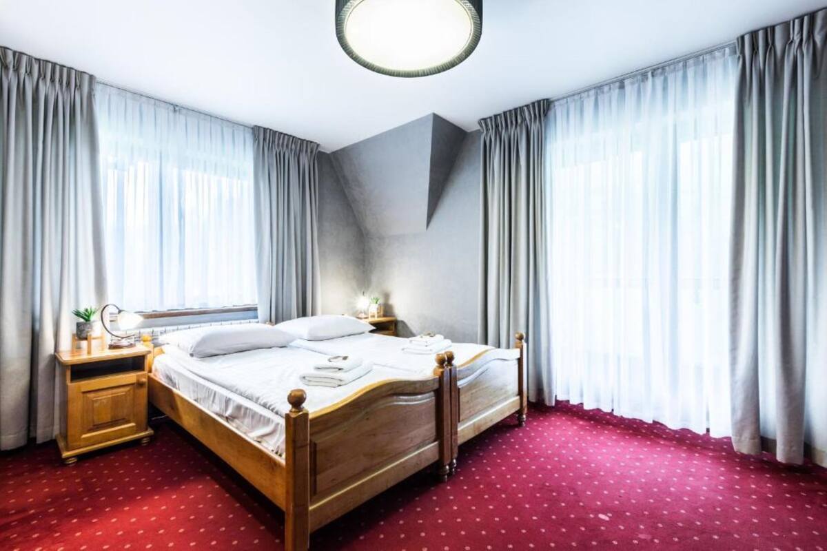 Una cama doble bien tendida con sábanas blancas, cabecera de madera y alfombra roja con lunares. Hay una mesita de noche con una lámpara y una pequeña planta. La habitación tiene ventanas grandes con cortinas translúcidas y un moderno aplique de techo.