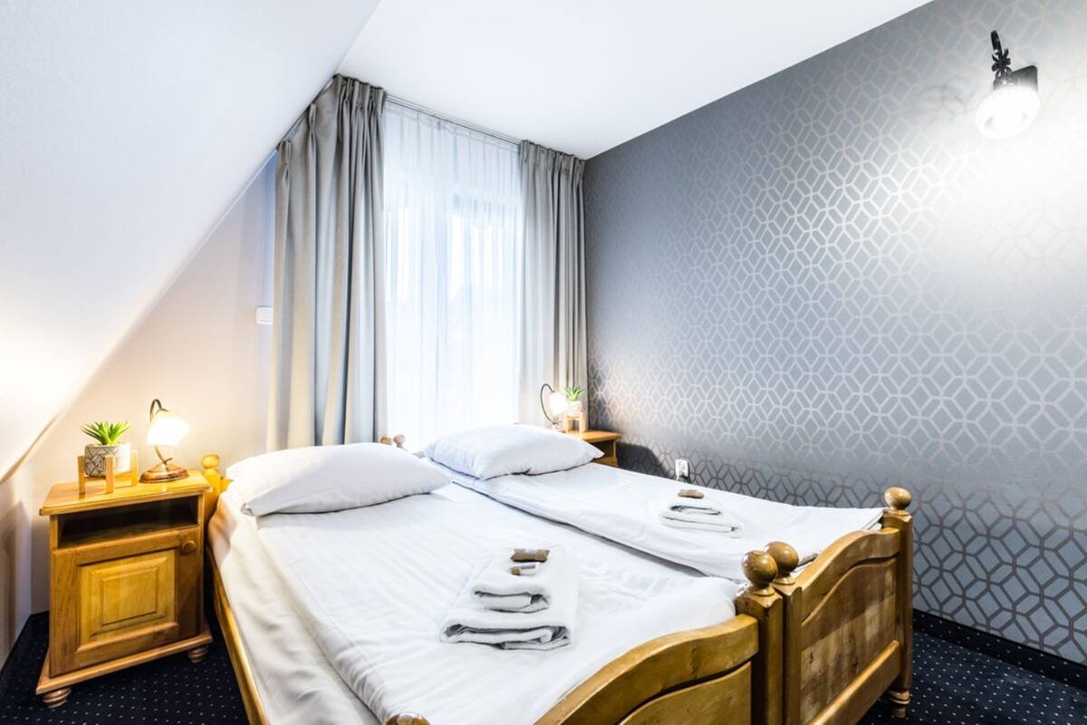 Habitación de hotel con dos camas, una mesita de noche con lámpara, una pequeña mesa con planta y una ventana con cortinas.