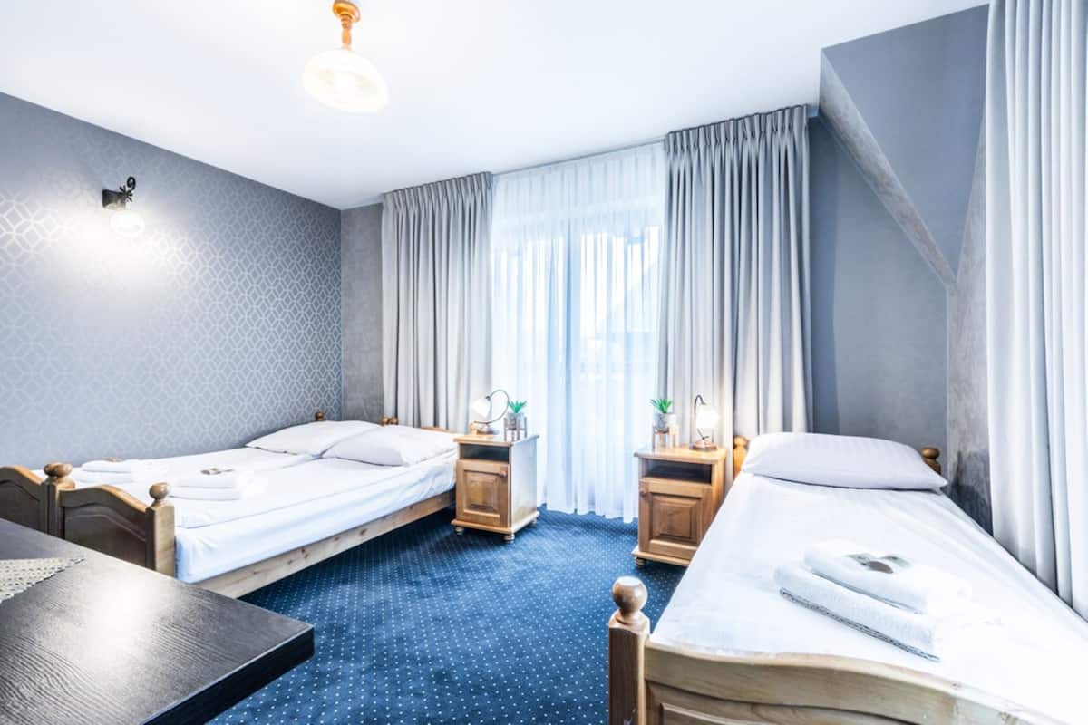 Habitación de hotel con dos camas individuales, una mesita de noche de madera con lámpara, una pequeña mesa y una ventana con cortinas.