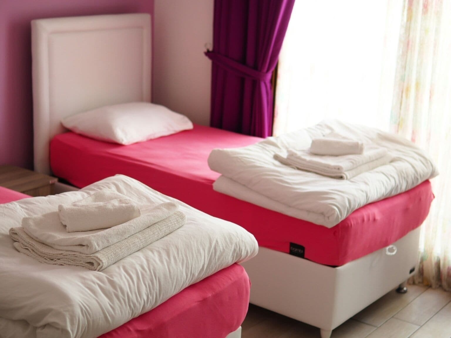 Duas camas com roupa de cama rosa e branca, uma cabeceira branca e uma cortina roxa.