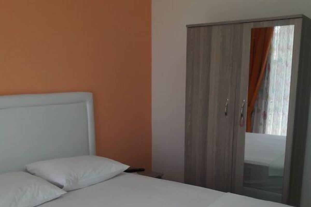 A neatly made bed with white linens and pillows, a grey wardrobe with a mirror, and a window with curtains.