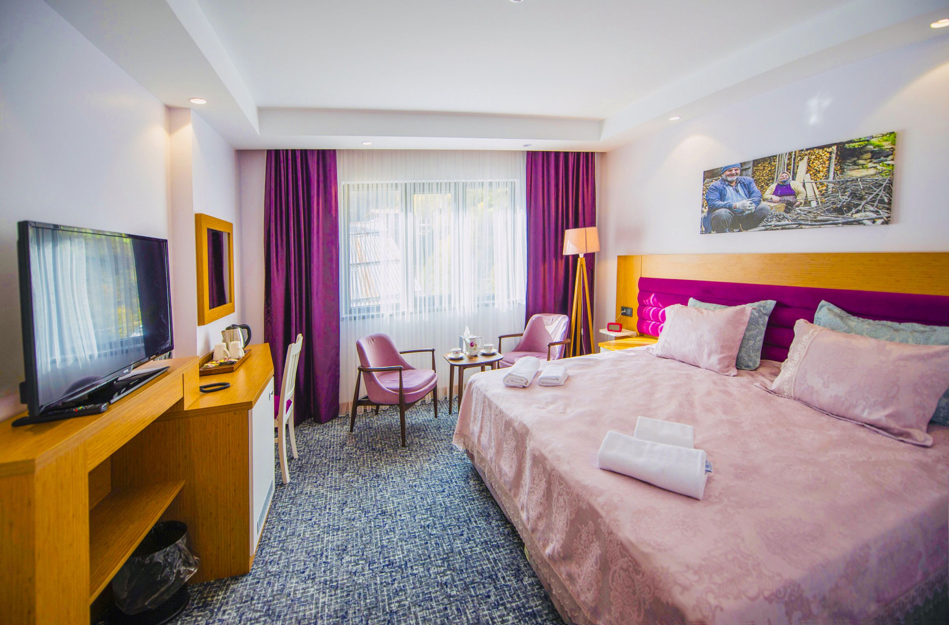 Una habitación de hotel con cama, escritorio con televisor, silla, lámpara y un cuadro en la pared.
