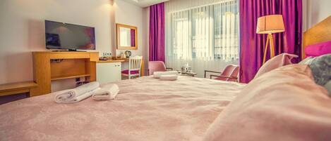 A hotel room with a large bed, a desk with a TV, a chair, a lamp, and a window with curtains.