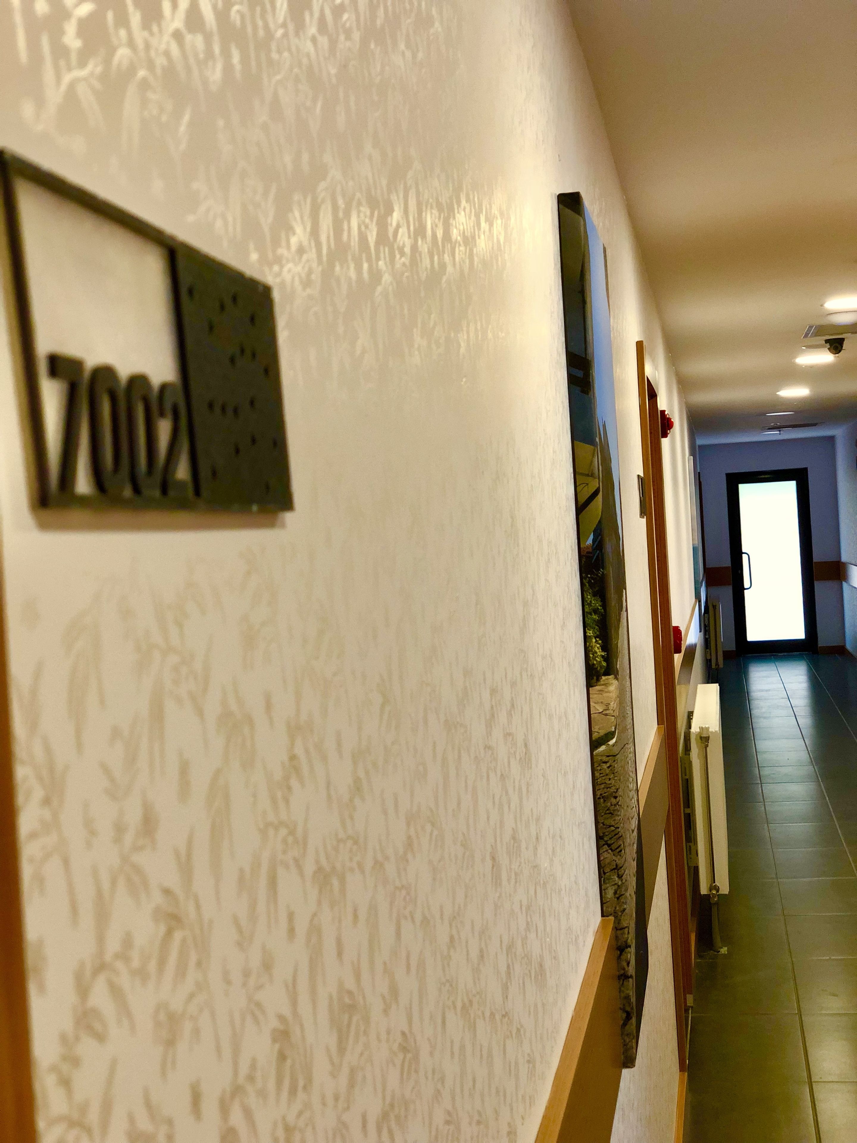 A hallway with a number sign '7002' on the wall, a patterned wallpaper, and a door at the end.