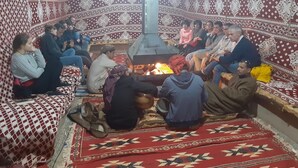 A group of people sitting around a central fire in a room with patterned walls and a checkered floor. - Wadi Rum Firecamp (Wadi Rum)