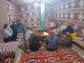 A group of people sitting around a central fire in a room with patterned walls and a checkered floor. - Wadi Rum Firecamp (Wadi Rum)