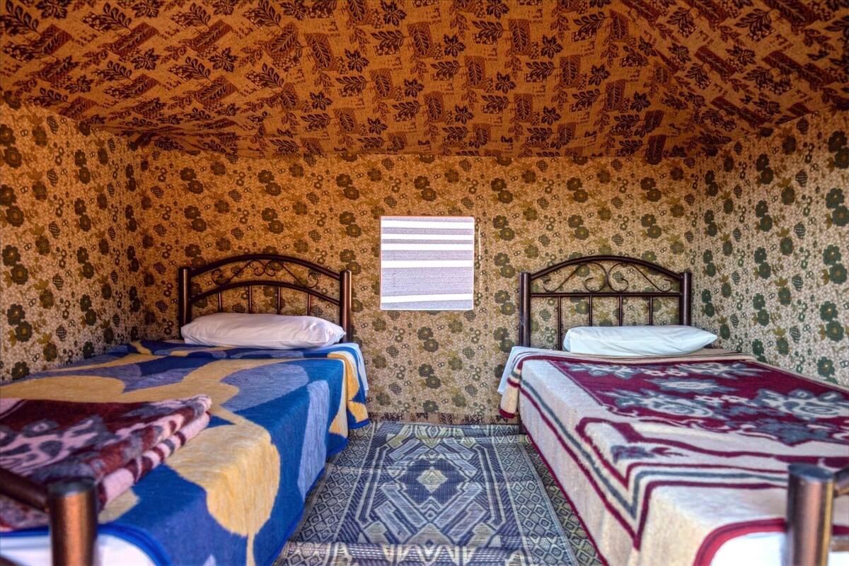 A room with two beds, patterned wallpaper, and a window with horizontal blinds.