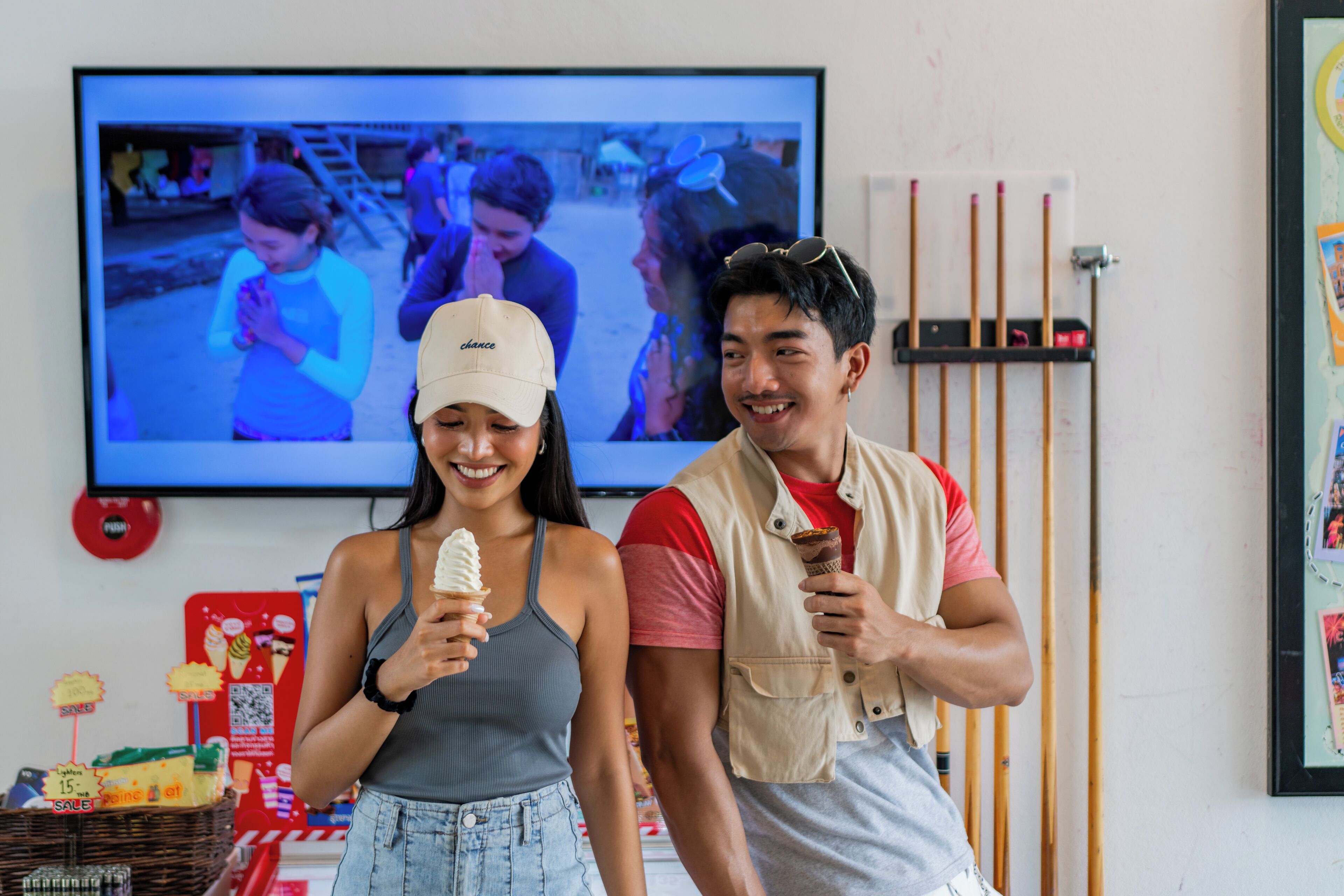 Two people holding ice cream cones in front of a television displaying a beach scene.