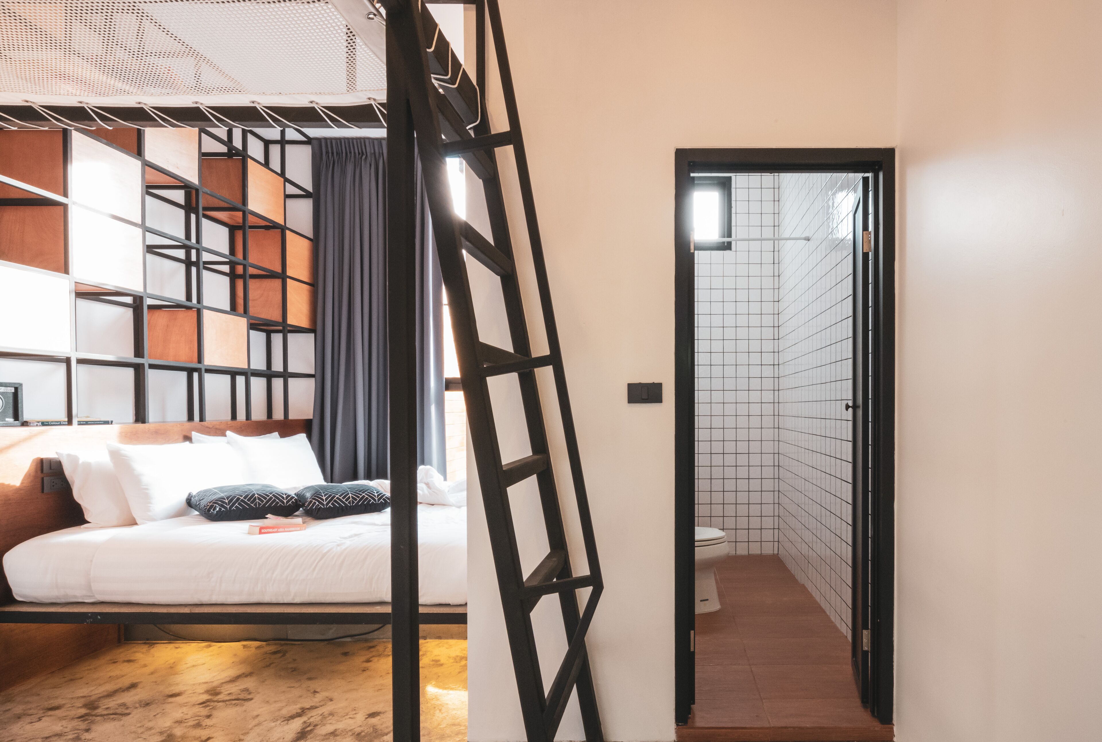A modern hotel room with a bed, a wooden headboard, a black metal ladder, and a view of the bathroom.
