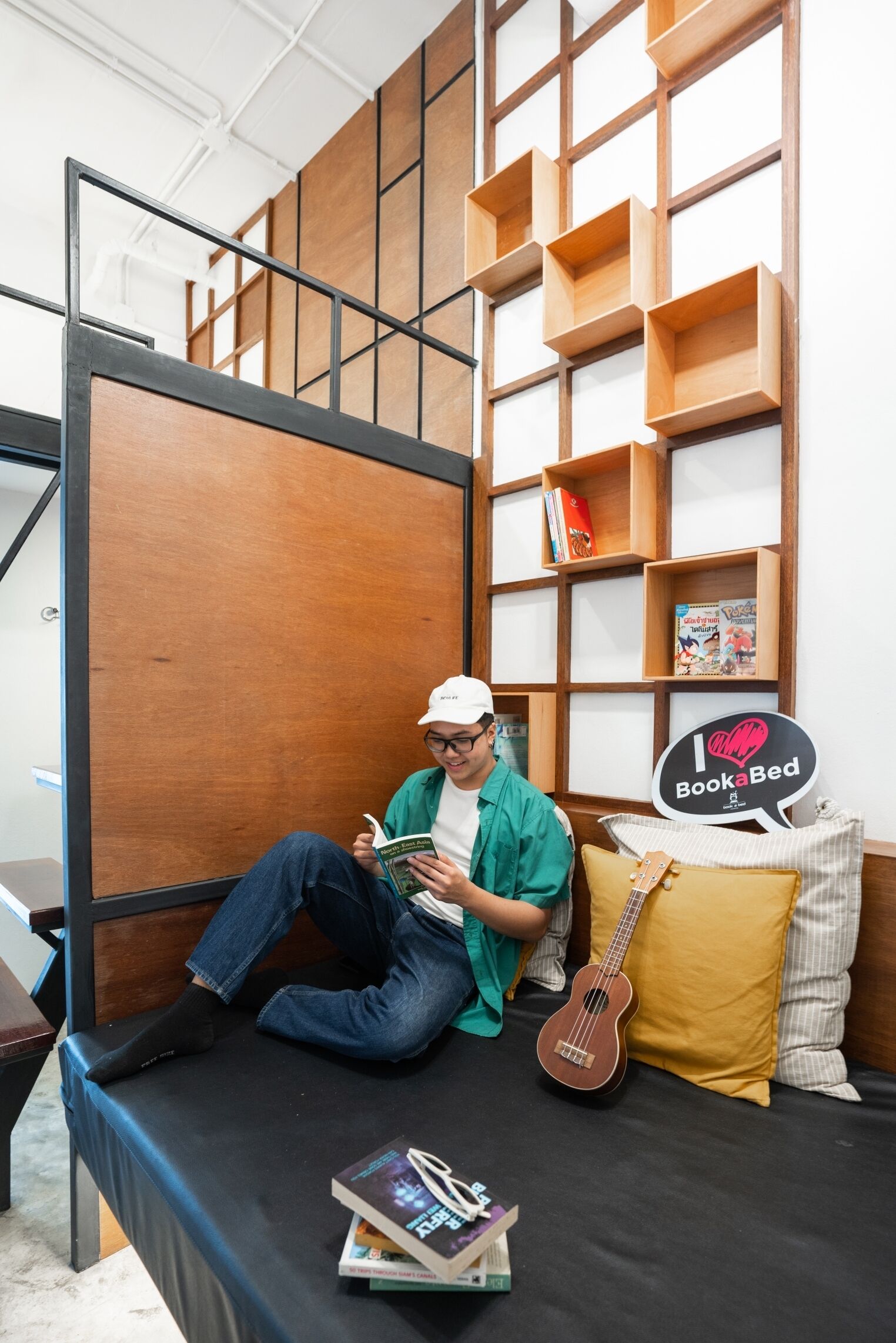 Eine Person sitzt auf einem Bett in einem Zimmer mit einem Bücherregal und einem Schild mit der Aufschrift 'I love BookBed'.
