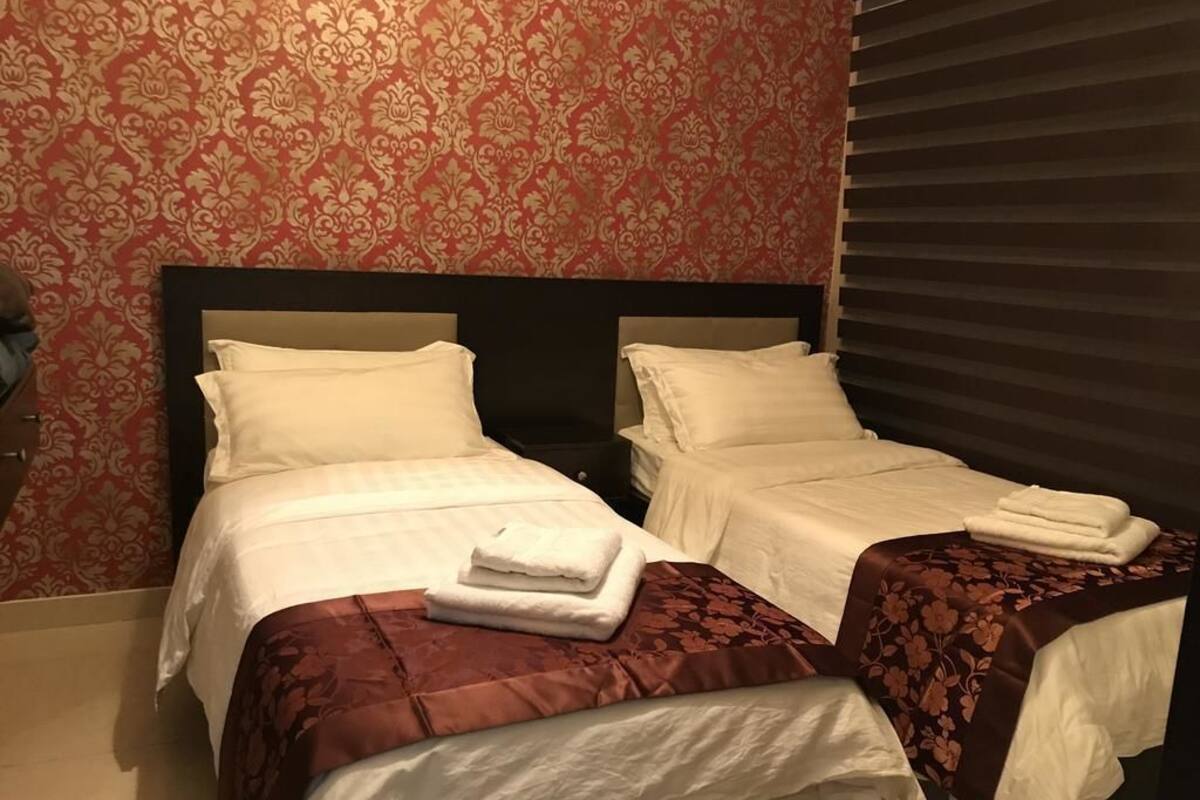 A hotel room with two beds, each with white linens and a brown bedspread. The room has a patterned wallpaper and a window with blinds.