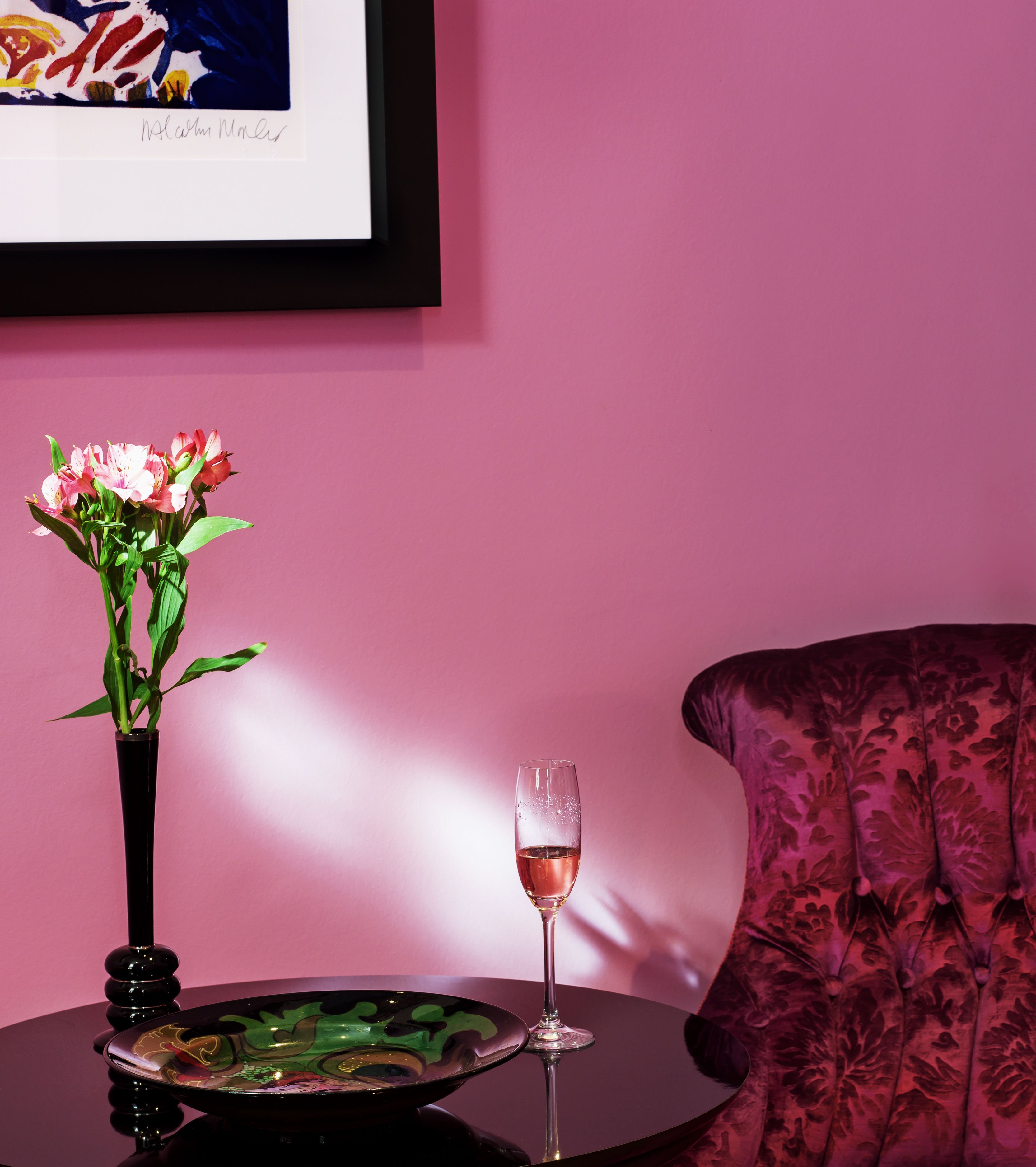 Uma mesa com um vaso de flores, um copo de vinho e um prato decorativo em frente a uma parede rosa.