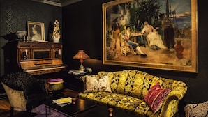 Una habitación con un sofá antiguo, un piano, un cuadro grande y una lámpara decorativa.